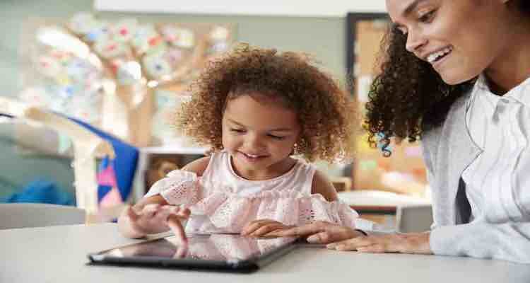 Young mixed race schoolgirl using a tablet computer with a female infant school teacher, working one on one together in a classroom, close up
